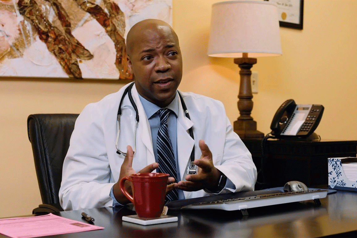 A doctor sitting behind a desk gestures with his hands as he speaks to someone out of frame.